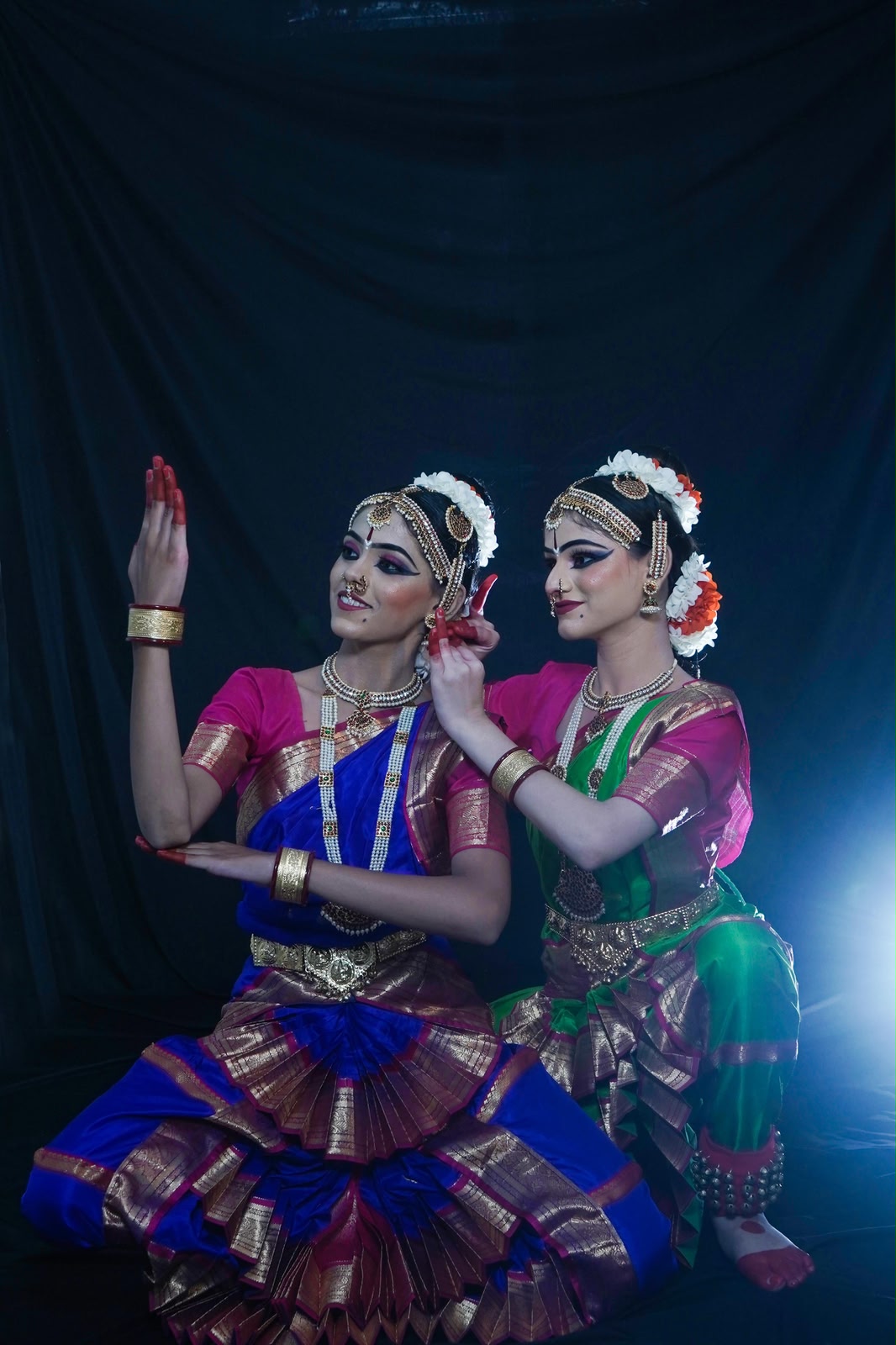 Dhruvi and Hetvi seated together against a dark draped backdrop, full Bharatanatyam costume with headpieces and necklaces, one hand raised in a mudra beside the other dancer’s face.